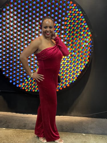 Reviewer posing in the burgundy Nightfall One Shoulder Velvet Jumpsuit in front of a colorful light display.