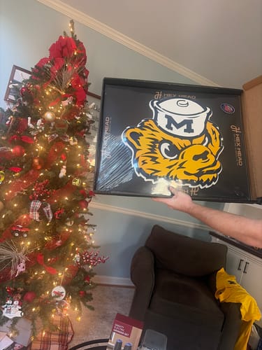 Customer holding the Michigan Wolverines wall art in its packaging in a home with a Christmas tree.