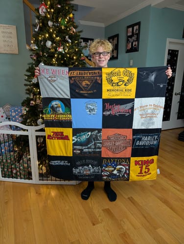 Reviewer holding a custom Premium T Shirt Quilt made from graphic shirts in front of a Christmas tree.