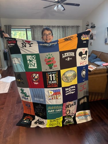 Customer's son smiling while holding up his large, colorful Premium T-Shirt Quilt in a living room.