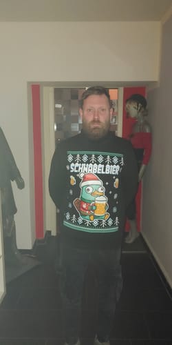 Ein Kunde trägt den schwarzen SCHNABELBIER-Pullover mit weihnachtlichem Motiv in einem Flur.