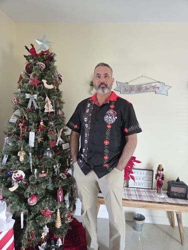 Reviewer wearing the black El Sancho Clause Guayabera shirt while standing next to a decorated Christmas tree.