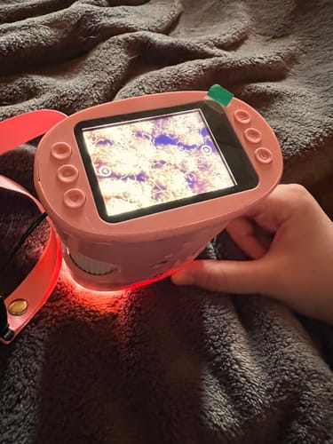 Ein Rezensent hält das rosa Edubini Abenteuer-Mikroskop, das am Boden rot leuchtet und eine vergrößerte Ansicht zeigt.