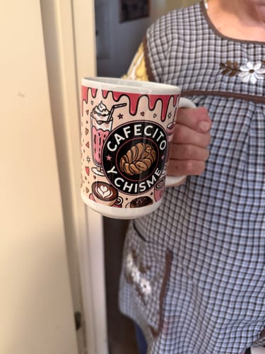 A person holding the Cafecito Y Chisme glossy mug, showing its colorful coffee-and-pastry-themed design.