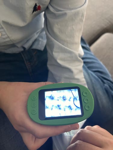 Ein Kunde hält das grüne Abenteuer-Mikroskop in der Hand, auf dessen Bildschirm eine stark vergrößerte Textur zu sehen ist.