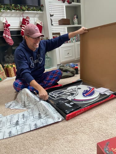 A customer smiles while unboxing the Buffalo Bills Helmet 3D metal wall art in a festive living room.