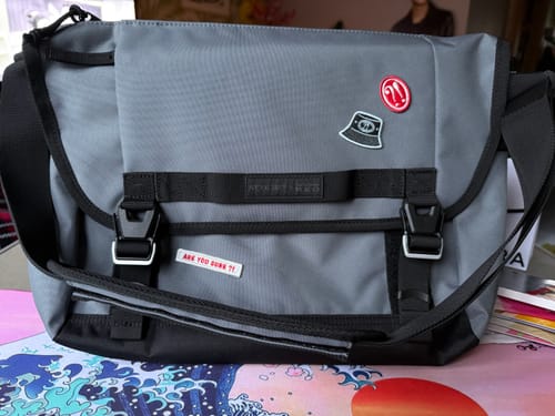 Customer's grey ARE YOU SURE X SAMSONITE messenger bag with black straps, buckles, and several decorative patches.