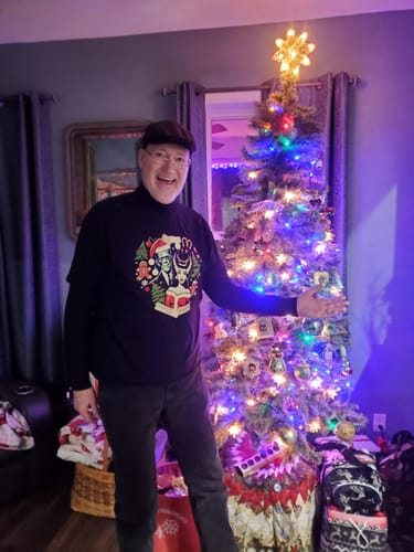 Happy reviewer wearing the Frankie & Liz Christmas Card T-Shirt, showing a great fit next to a Christmas tree.