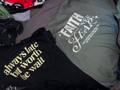 Reviewer's two new shirts laid out, showing the back graphics 'FAITH OVER FEAR' and 'always late but worth the wait'.