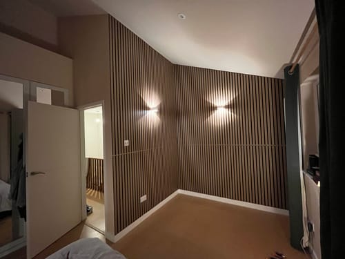 Customer's bedroom with natural oak slat wall panels installed on corner walls, illuminated by two modern wall lights.