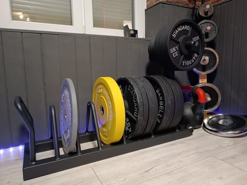 Rezensent zeigt die schwarze Armored Tusker Hantelscheibenablage, die in einem Heimstudio mit Hantelscheiben beladen ist.