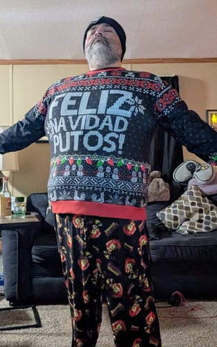 Reviewer standing with arms outstretched while wearing the black FELIZ NAVIDAD PUTOS Sweatshirt in a living room.
