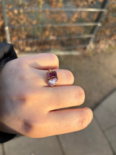 Customer's hand wearing the elegant Leesha Birthstone Ring with a red and light purple stone outdoors.