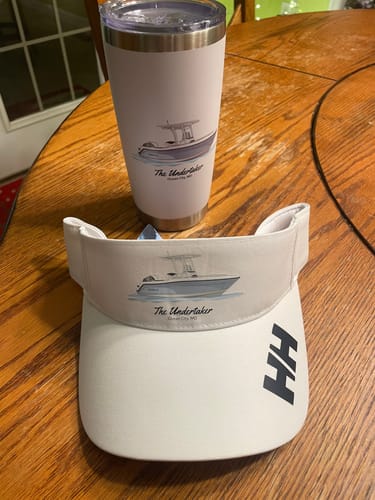 Customer's custom boat artwork on a white tumbler and matching visor displayed on a wooden table.