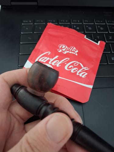 Customer holding the red packaging for Cartel Cola THCA Flower and a dark wooden pipe over a laptop keyboard.