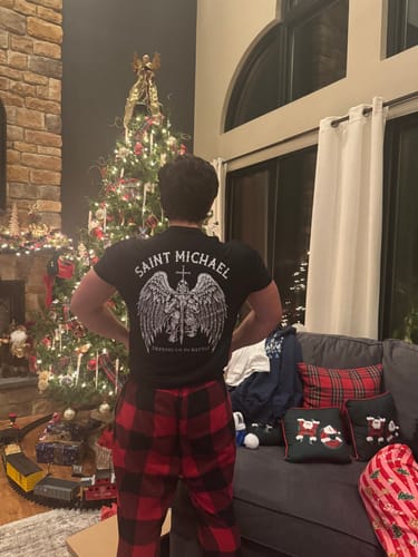 Reviewer showing the back graphic of the black Saint Michaels Tee in a living room.