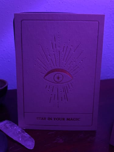 Reviewer's Stay Golden postcard with a foil eye design, displayed on a wooden surface under purple light.