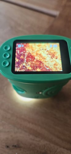 Rezensent zeigt das eingeschaltete, grüne Edubini Abenteuer-Mikroskop, dessen Bildschirm eine stark vergrößerte Ansicht zeigt.