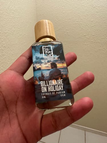 Reviewer holding the glass bottle of Billionaire On Holiday perfume, which has a wood-grain cap and a scenic label.