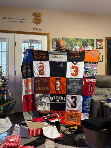 Customer holding a large Premium T Shirt Quilt made from sports and Harley-Davidson shirts, with a Christmas tree in the background.