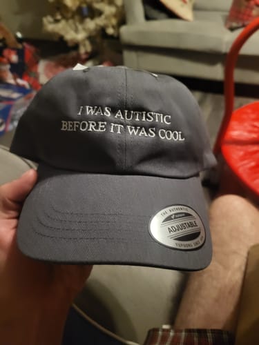 Customer holding a grey 'Autistic Before It Was Cool' dad hat, showing the white embroidered text.