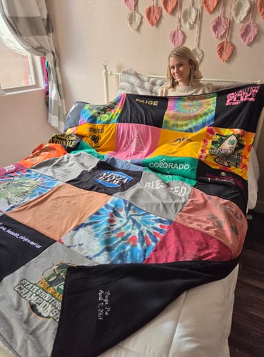 Customer sitting on a bed, displaying a large colorful Premium T-Shirt Quilt made of many different shirts.
