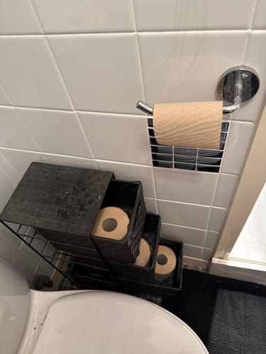 Customer's Unbleached Bamboo Toilet Paper on a holder, with extra rolls stored in a black bathroom organizer.