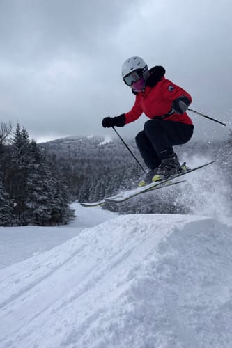 Reviewer wearing the red ski jacket with a black faux fur hood while getting air on skis.