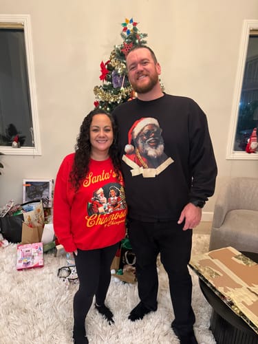 Reviewer wearing the black Grilled Up OG Santa Christmas Sweatshirt in front of a Christmas tree with another person.