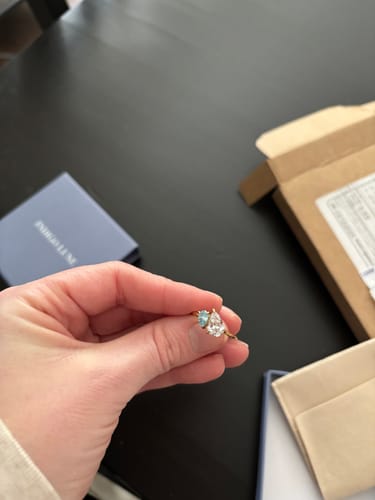 Customer holding the Leesha Birthstone Ring, showing a light blue and a clear pear-cut stone on a gold band.