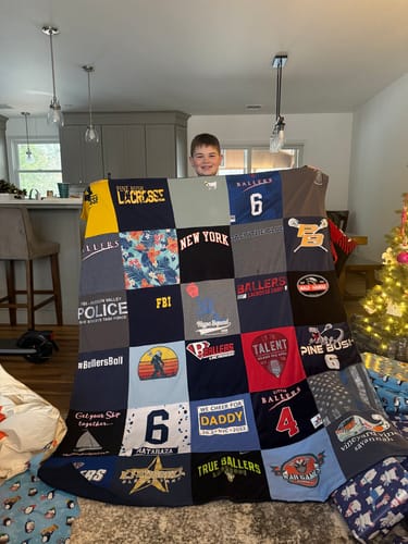 A customer's son smiles while holding up a large, multi-panel Premium T Shirt Quilt in a living room.