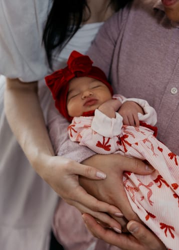 Customer's newborn baby wearing the Christmas Bow Take-Me-Home Set with its printed gown and red bow headband.