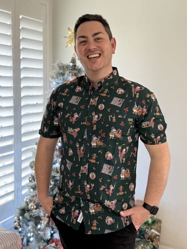 Customer smiling while wearing the green 'The Storytellers' short sleeve shirt in front of a Christmas tree.