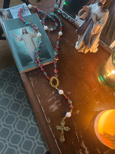 Reviewer's red-blue glass bead rosary with its ornate cross laid out on a table beside prayer items.
