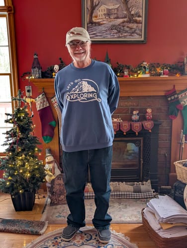 Customer smiling while wearing the blue Lets Go Exploring Sweatshirt in front of a fireplace.