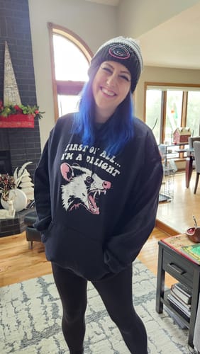 A reviewer smiling while wearing the black "First Of All I'm A Delight Hoodie" in a living room.