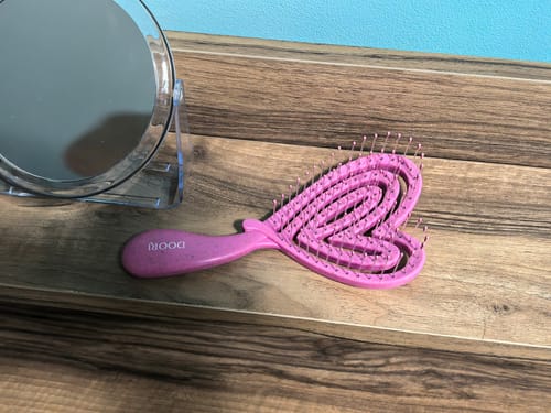 Рецензент показва своята розова, сърцевидна четка за коса Doori на дървен плот до огледало.