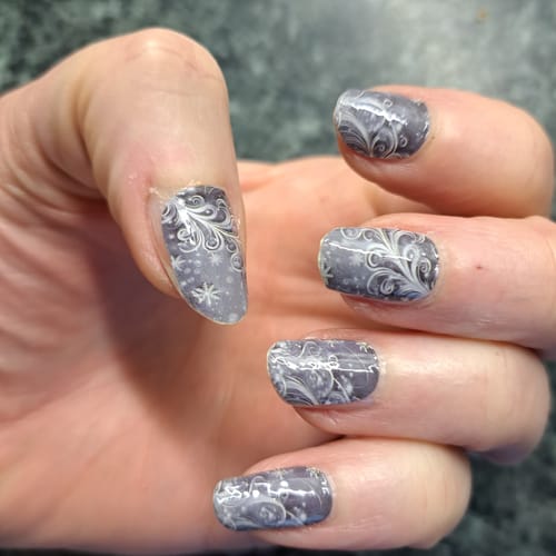 Nahaufnahme einer Hand, die eine Maniküre mit den grauen 'Grey Frost'-Nagelfolien mit elegantem, silbernem Wirbelmuster zeigt.