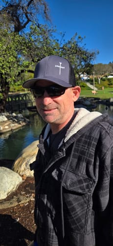 Customer wearing the grey and black Nail Cross Lower Left Hat with a white cross logo outdoors.