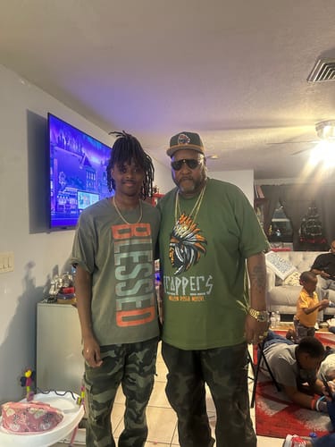 A reviewer wearing the olive green Trappers t-shirt with a graphic, posing indoors with another person.