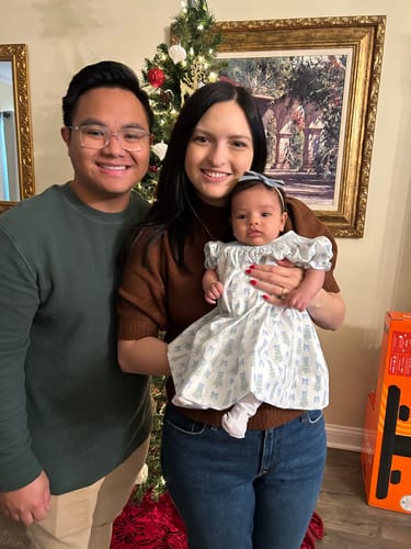 A reviewer holds their baby, who is wearing the light blue smocked bishop dress in front of a Christmas tree.