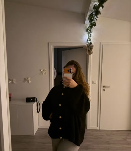 Rezensentin trägt den schwarzen Oversized Cardigan 'CozyGlam' mit goldenen Knöpfen in einer Spiegelaufnahme.