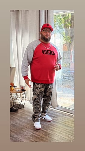 Reviewer standing indoors, wearing a red sweatshirt with grey sleeves and camouflage pants.