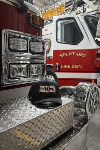 Customer's black SNAPBACK HOSE HAT with a 'Station 10' patch resting on the bumper of a red fire truck.