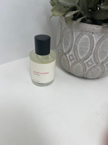 Customer's Sunset in Paris perfume bottle on a white surface next to a decorative plant.