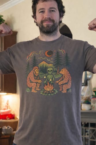 Reviewer wearing the dark grey Campfire Friends T-Shirt, showing the fit and graphic of Bigfoot, an alien, and a bear at a campfire.