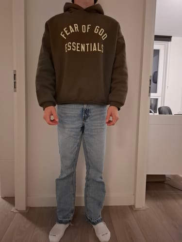 Klant toont de pasvorm en militaire kleur van de Fear of God Essentials hoodie.