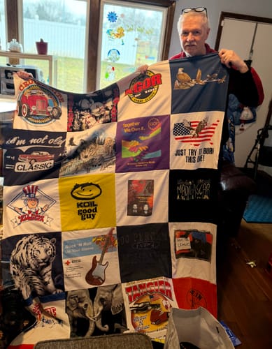 A smiling person holds up a large, colorful Premium T Shirt Quilt made from many different t-shirt squares.