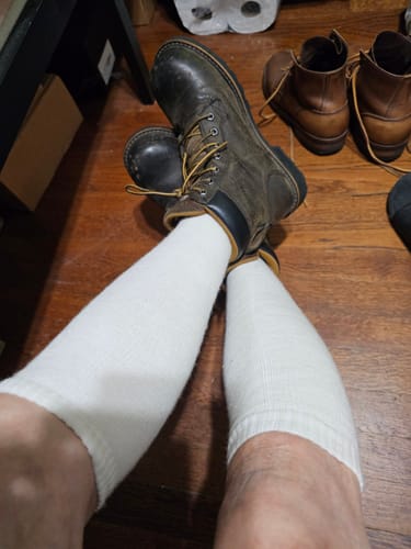 Customer sitting on a floor wearing long, white Men's Alpaca Socks with a dark work boot.