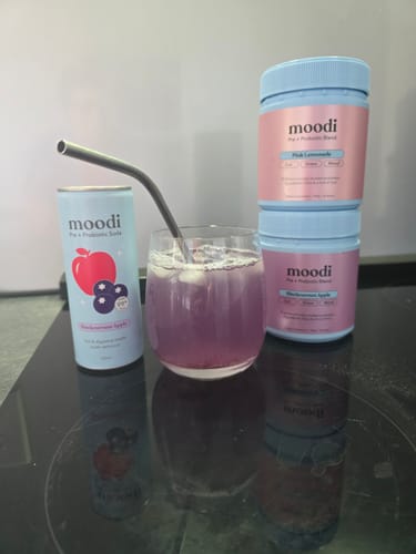Customer's prepared Moodi Blackcurrant Apple drink in a glass, shown with the can and powder tubs.